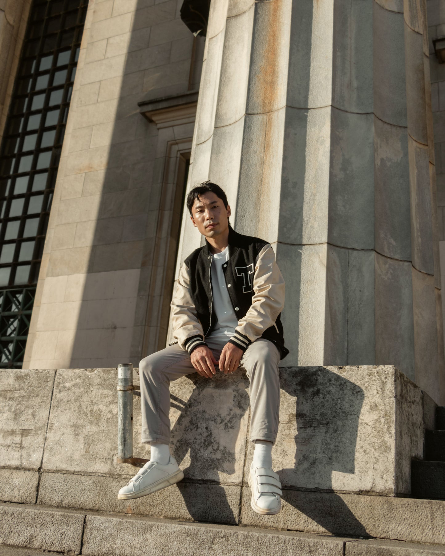 Male model wearing Tamp Coffee's black and cream varsity jacket made of leather and wool, with cream stripes on the wrists and waist. The model is sitting on a large step of a traditional building with Romanesque columns