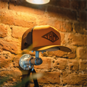 A vibrant Orange Trucker Cap featuring an embroidered Tamp Coffee logo, suspended on a metal support, against a textured brick wall backdrop