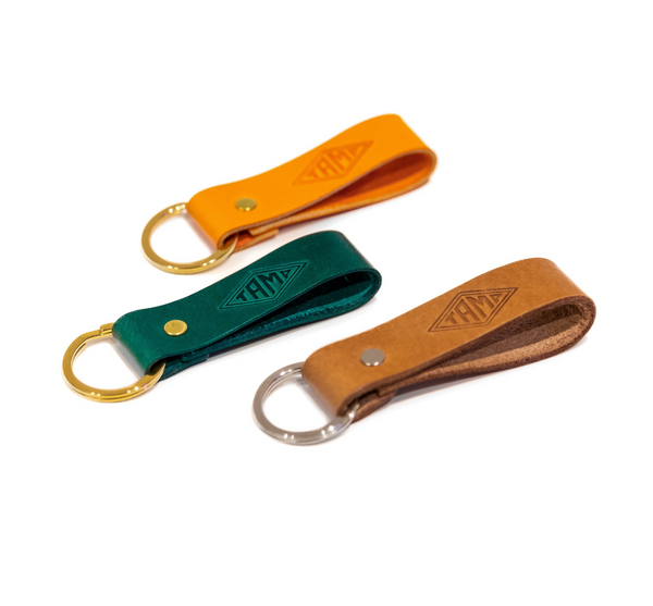 three leather keyrings in green, orange, and brown, handcrafted by Tamp Coffee. Each keyring showcases intricate stitching and a beautifully embossed logo, combining elegance and durability